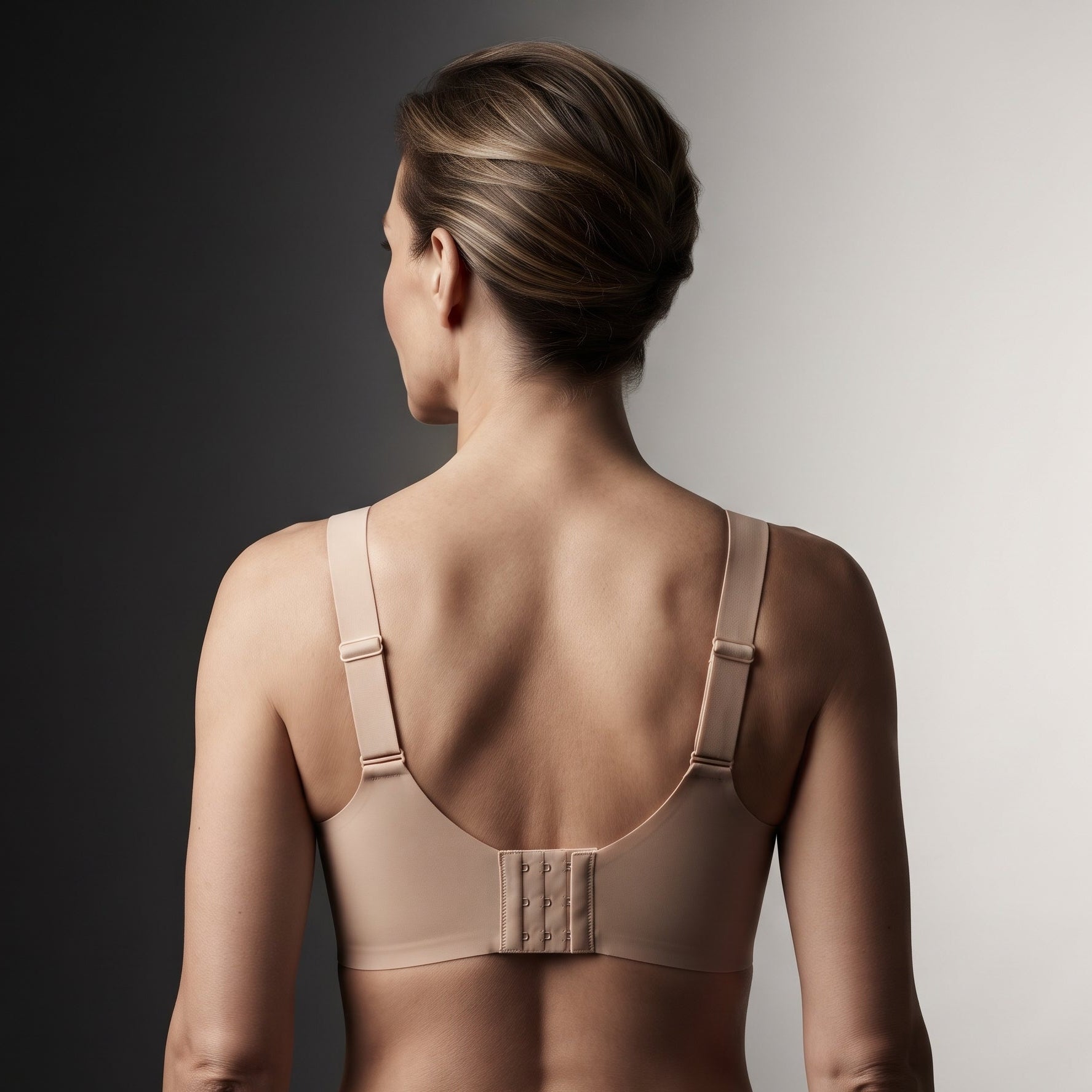 A woman shown from the back wearing a beige wireless bra with adjustable shoulder straps and a central hook-and-eye closure, photographed against a dark-to-light gray gradient studio background.