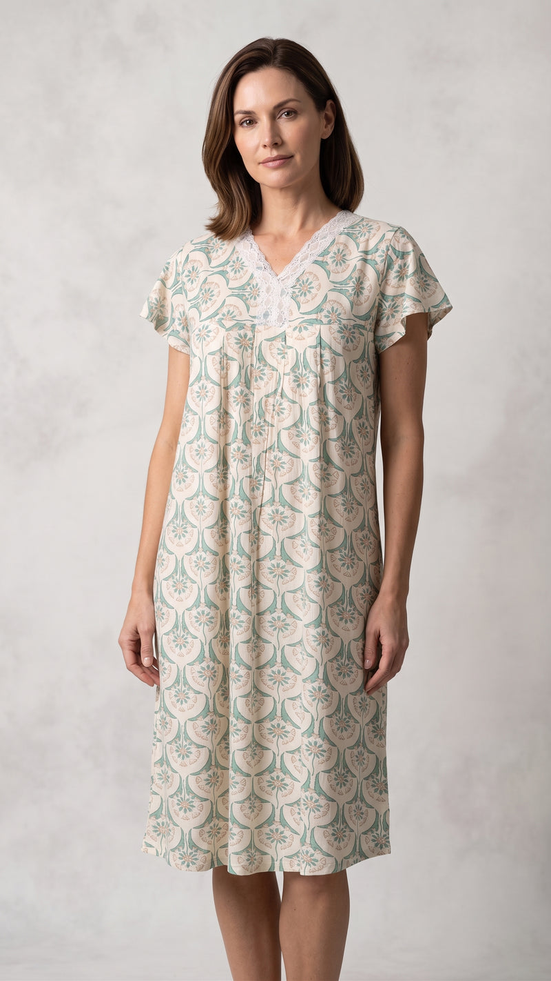 A woman modeling a short-sleeve V-neck nightgown with a beige and sage green floral block print pattern, set against a neutral textured gray background.