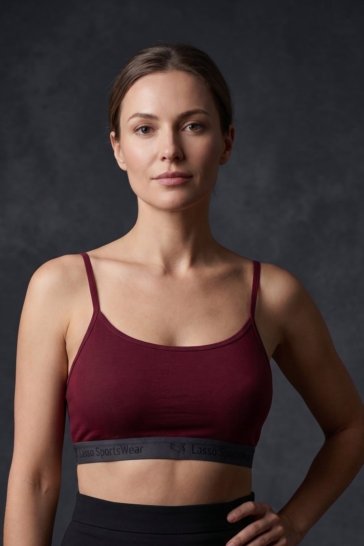 A woman modeling a dark burgundy sports bra with a dark gray underband and black high-waisted skirt against a dark gray studio background.