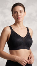 A woman modeling a black underwire bra with subtle floral lace detailing and sheer mesh panels, set against a textured light gray background.