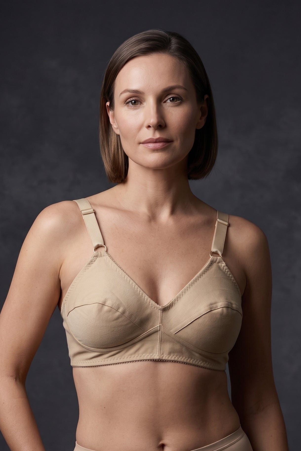 A woman modeling a beige nursing bra and matching high-waisted underwear against a dark gray studio background.
