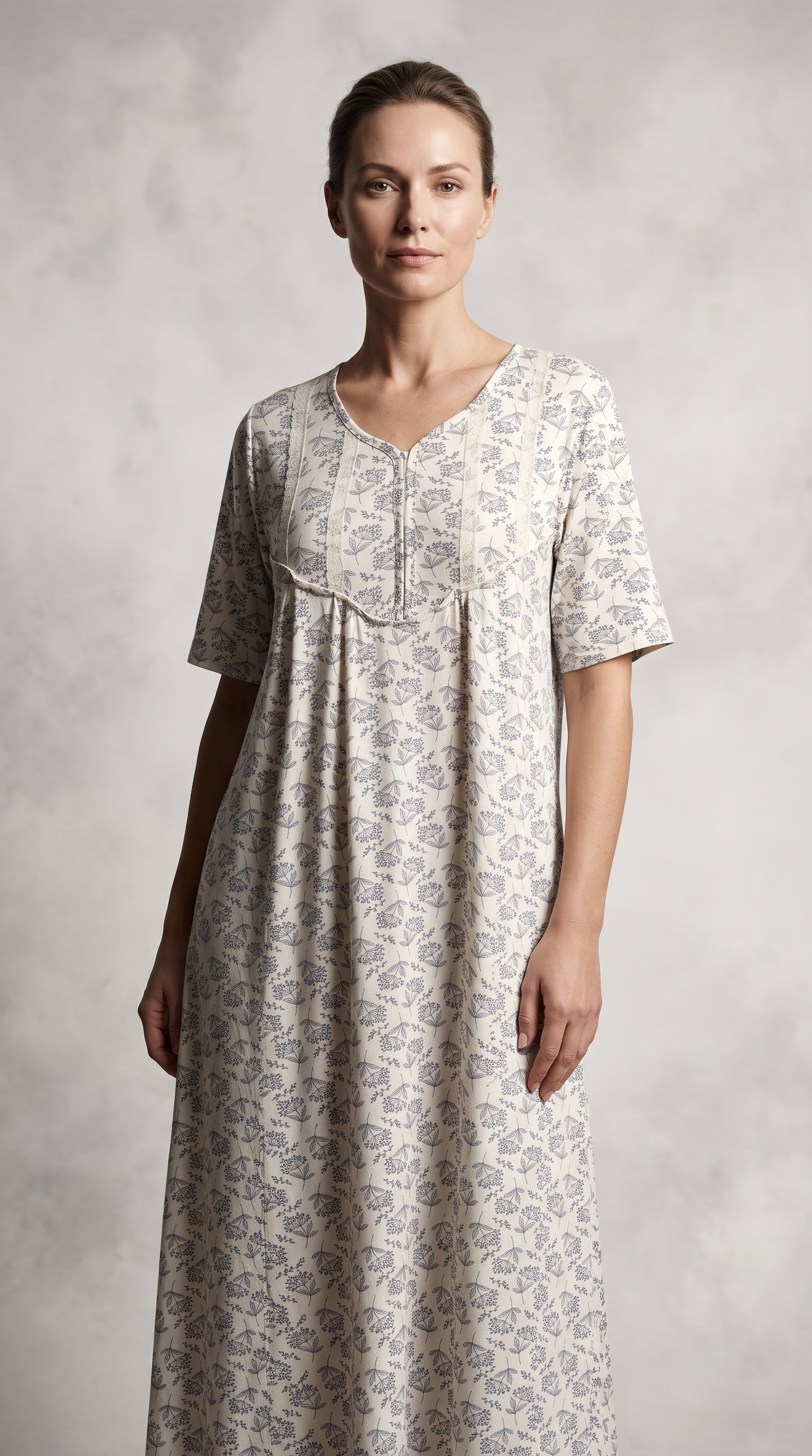 A woman modeling a short-sleeved, V-neck nightgown with a delicate gray floral block print on a warm beige fabric, set against a textured gray studio backdrop.