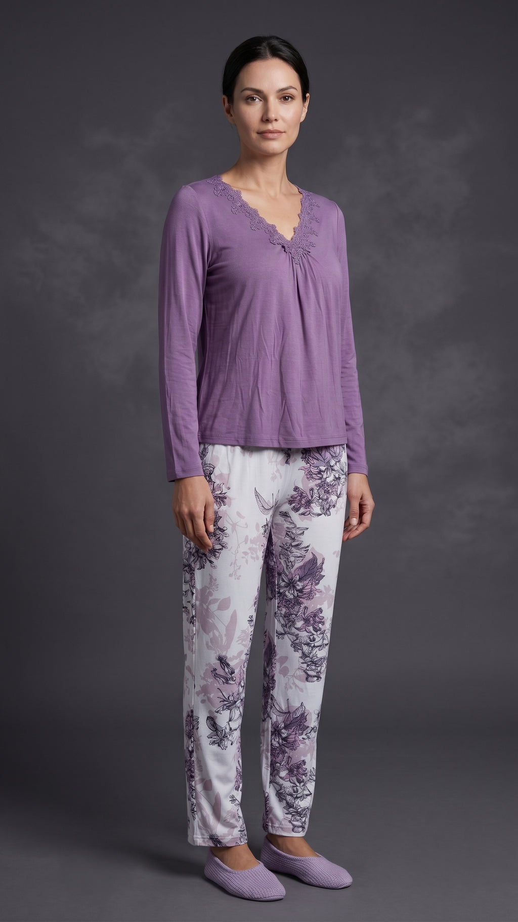A woman modeling a two-piece pajama set consisting of a lavender long-sleeve top with a gathered neckline and matching floral-print pants on a dark gray studio background.