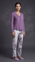 A woman modeling a two-piece pajama set consisting of a lavender long-sleeve top with a gathered neckline and matching floral-print pants on a dark gray studio background.