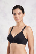 A woman modeling a black two-piece lingerie set consisting of a bralette with crossover straps and matching high-cut briefs, set against a light gray textured studio background.