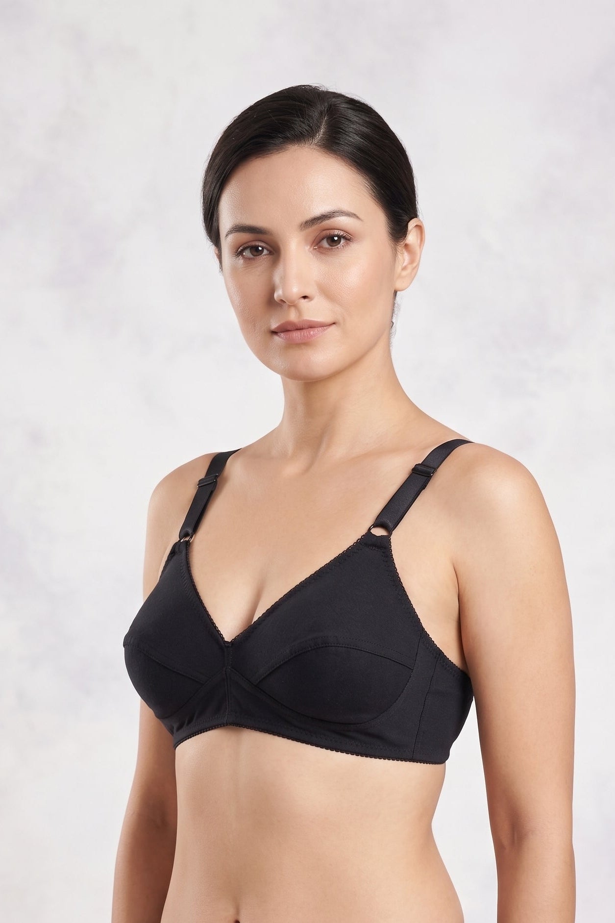 A woman modeling a black two-piece lingerie set consisting of a bralette with crossover straps and matching high-cut briefs, set against a light gray textured studio background.
