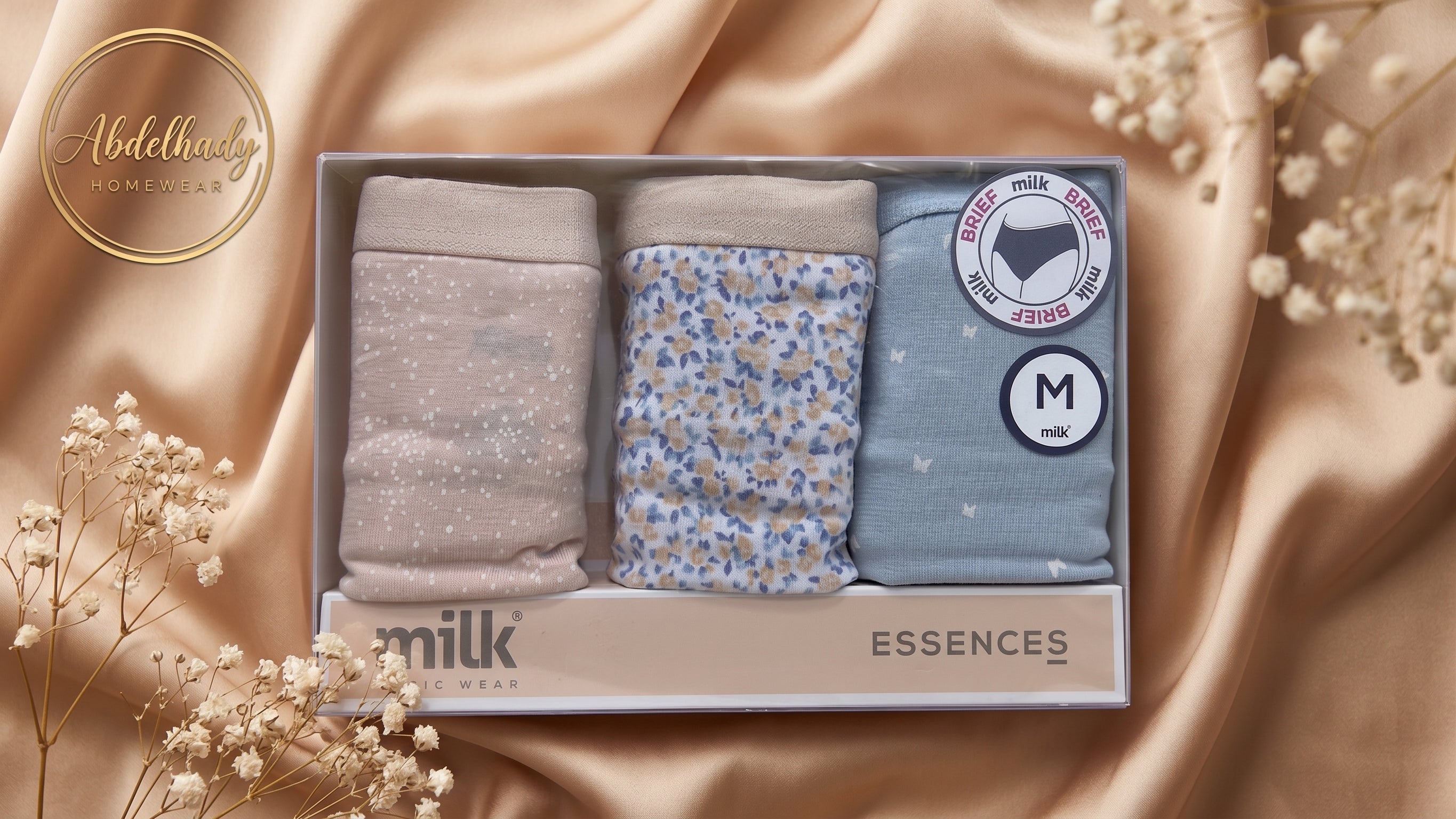 A boxed set of three pairs of women's underwear displayed in a gift box on draped champagne satin with baby's breath flowers. The set includes a light taupe glitter pair, a light blue floral print pair, and a light blue star print pair.