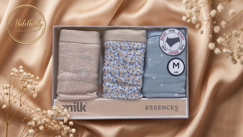 A boxed set of three pairs of women's underwear displayed in a gift box on draped champagne satin with baby's breath flowers. The set includes a light taupe glitter pair, a light blue floral print pair, and a light blue star print pair.