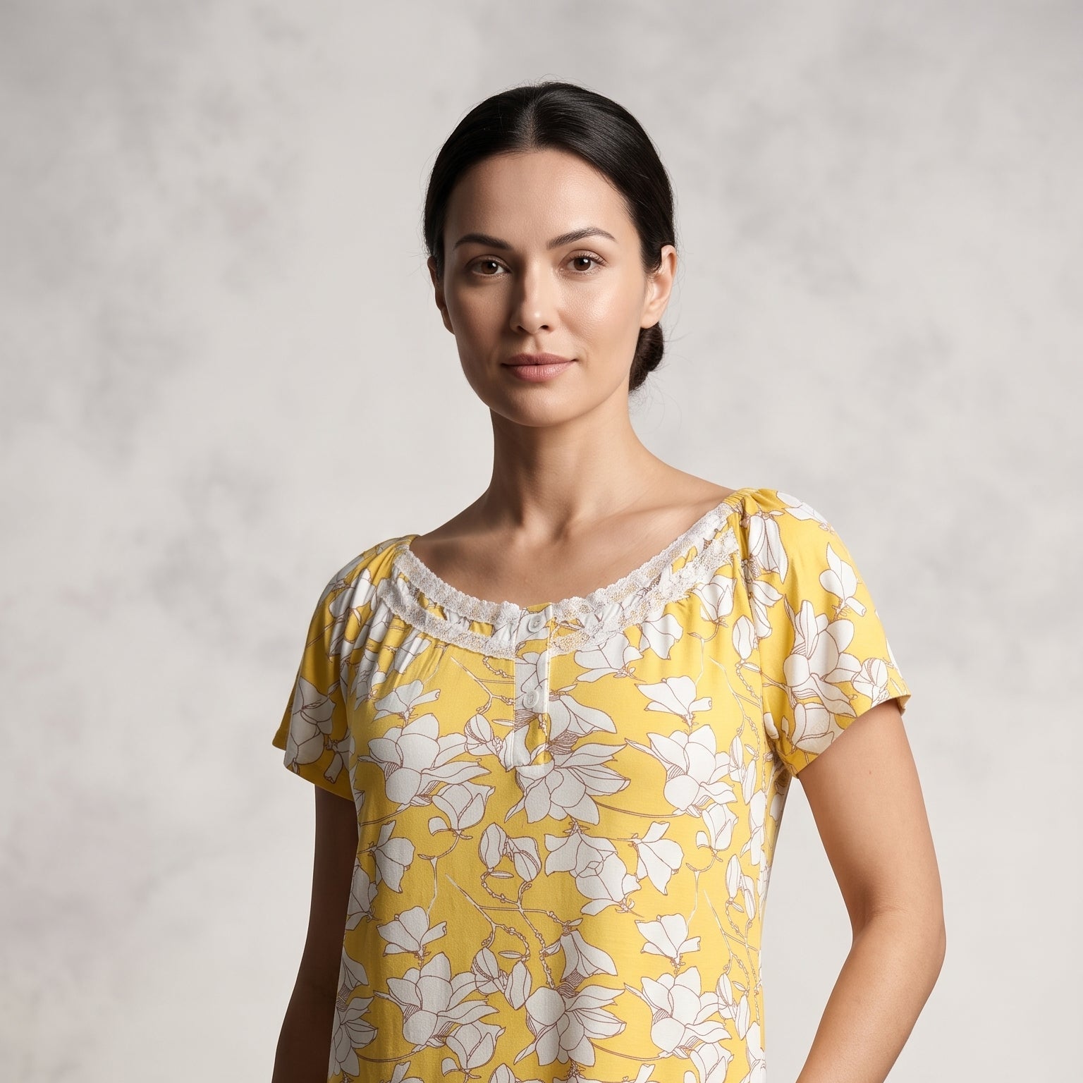 A woman modeling a short-sleeve yellow nightgown with a white floral print and a gathered neckline, set against a textured light gray background.