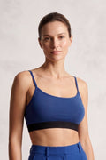A woman modeling a royal blue sports bra with a black band and matching blue high-waisted trousers against a textured light gray background.