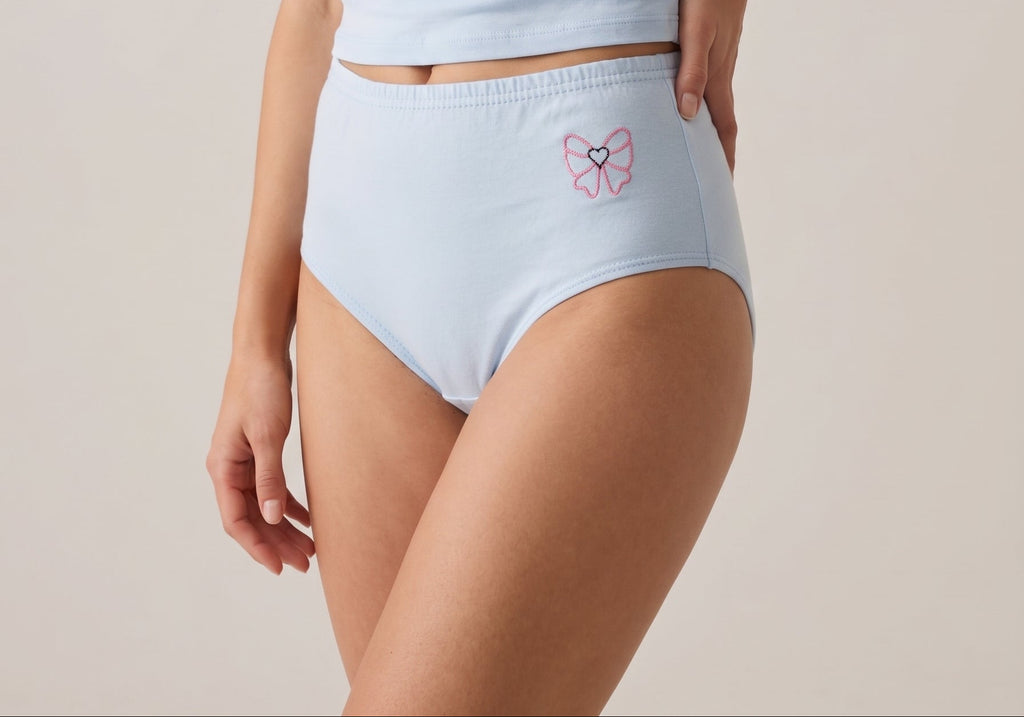 A model wearing a light blue short-sleeve crop top and matching high-waisted light blue underwear with a small pink butterfly graphic on the front, set against a plain beige studio background.