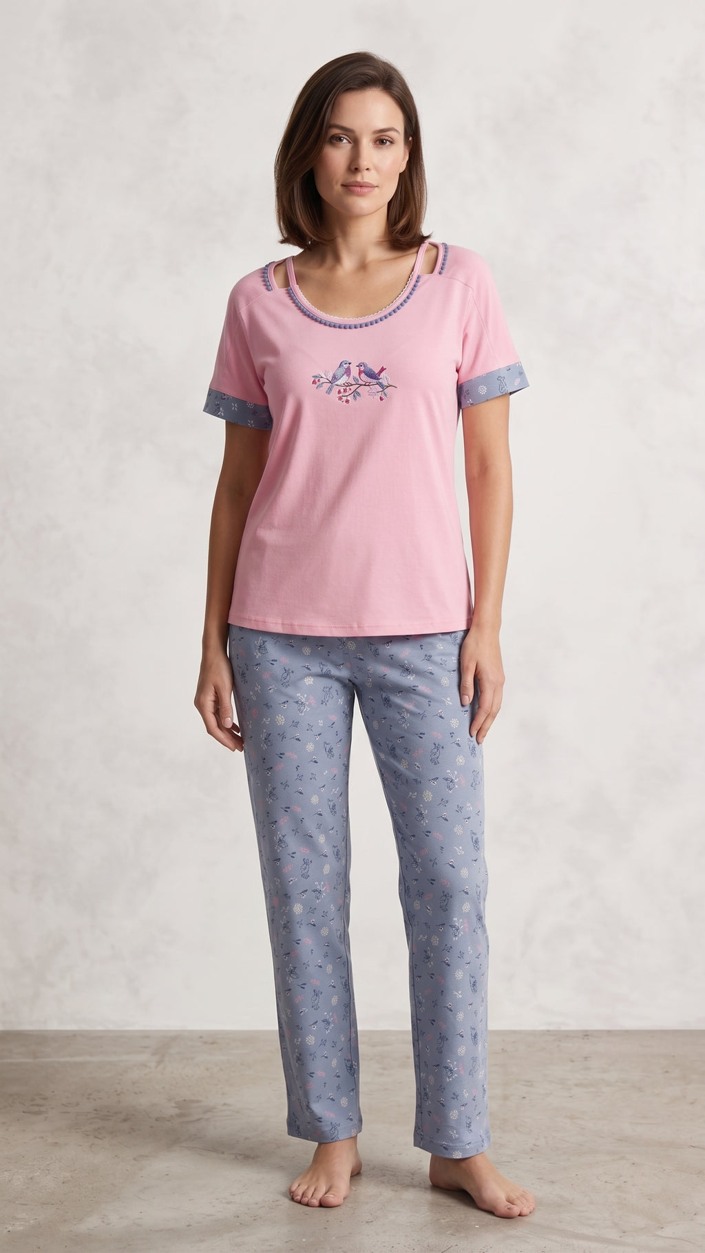 A woman modeling a pink short-sleeve pajama top with a small unicorn graphic on the chest and light blue patterned pajama pants, standing against a textured light gray wall.