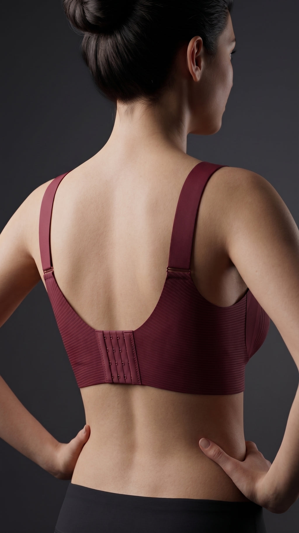 Rear view of a woman wearing a burgundy wireless sports bra with a structured back panel and wide shoulder straps, set against a dark gray studio background.